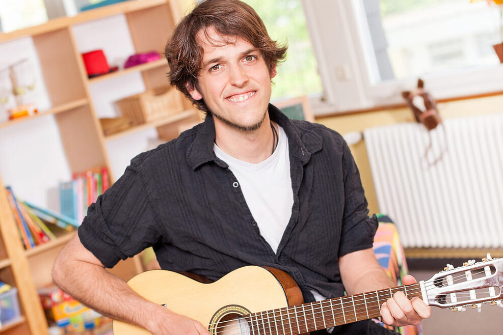 Ein junger Mann mit schwarzem Hemd hält eine braune Gitarre in der Hand und lächelt verschmitzt in die Kamera.
