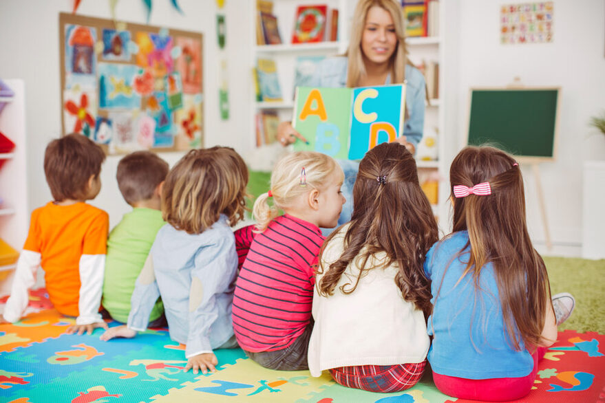 Sechs Kinder sitzen in einer Kita mit dem Rücken zur Kamera auf dem Boden vor einer Frau, die ein Bild mit Buchstaben in die Luft hebt