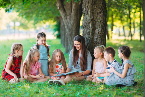 Eine Erzieherin lehnt an einem Baum und liest Jungen und Mädchen im Park ein Buch vor.