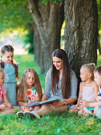 Eine Erzieherin lehnt an einem Baum und liest Jungen und Mädchen im Park ein Buch vor.