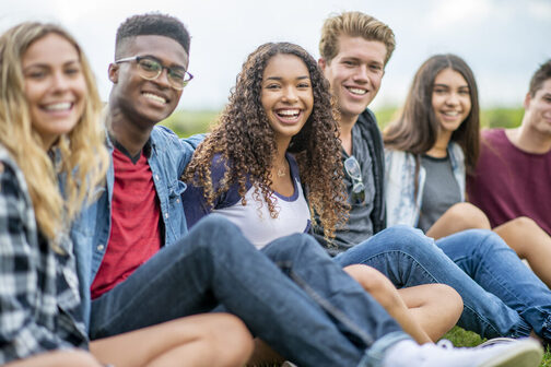 Eine Gruppe von sechs jungen Personen sitzt Arm in Arm zusammen auf einer Wiese und lächelt in die Kamera