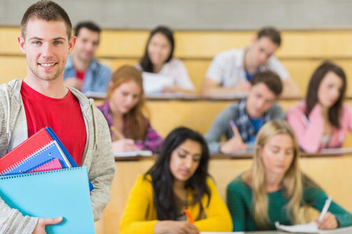 Ein lächelnder Student steht im Hörsaal und hält Unterlagen in der Hand, im Hintergrund sind andere Studenten zu sehen