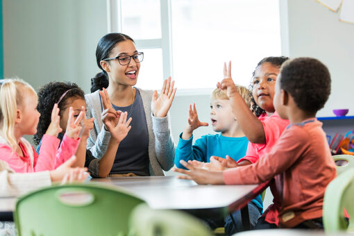 Eine junge Frau mit Brille sitzt mit sechs Kindergartenkindern an einem Tisch. Sie halten alle ihre Hände in die Luft.