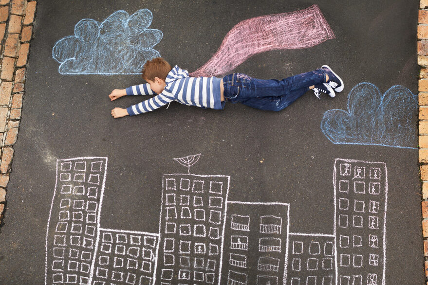 Ein kleiner Junge mit blau-weiß-gestreiftem Pullover liegt auf der Straße über einer mit Kreide gezeichneten Stadt